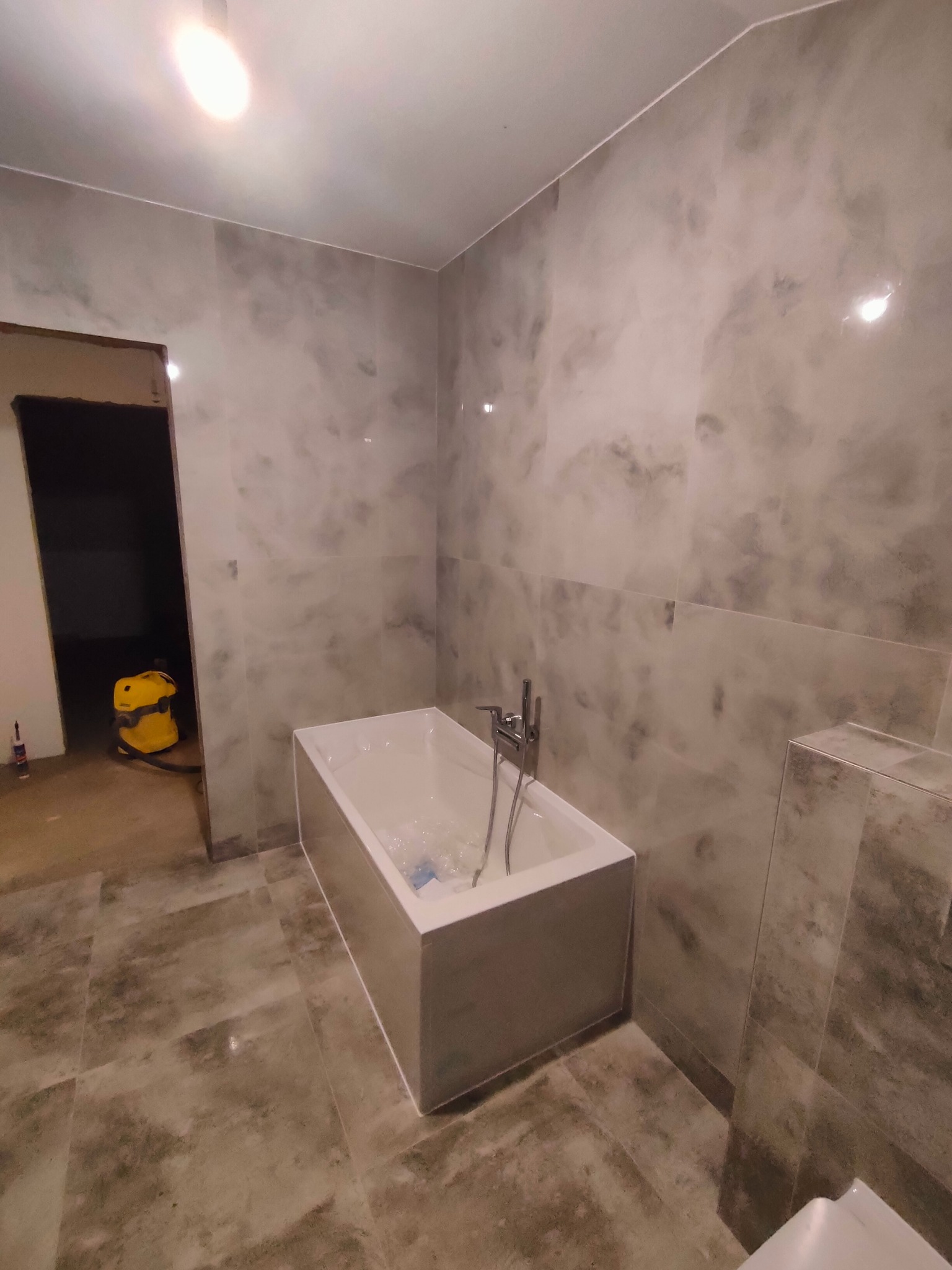 Wykończona łazienka w odcieniach szarości w Mińsku Mazowieckim, z wanną wypełnioną wodą i widocznym odkurzaczem w sąsiednim pomieszczeniu, sugerującym trwające prace porządkowe.