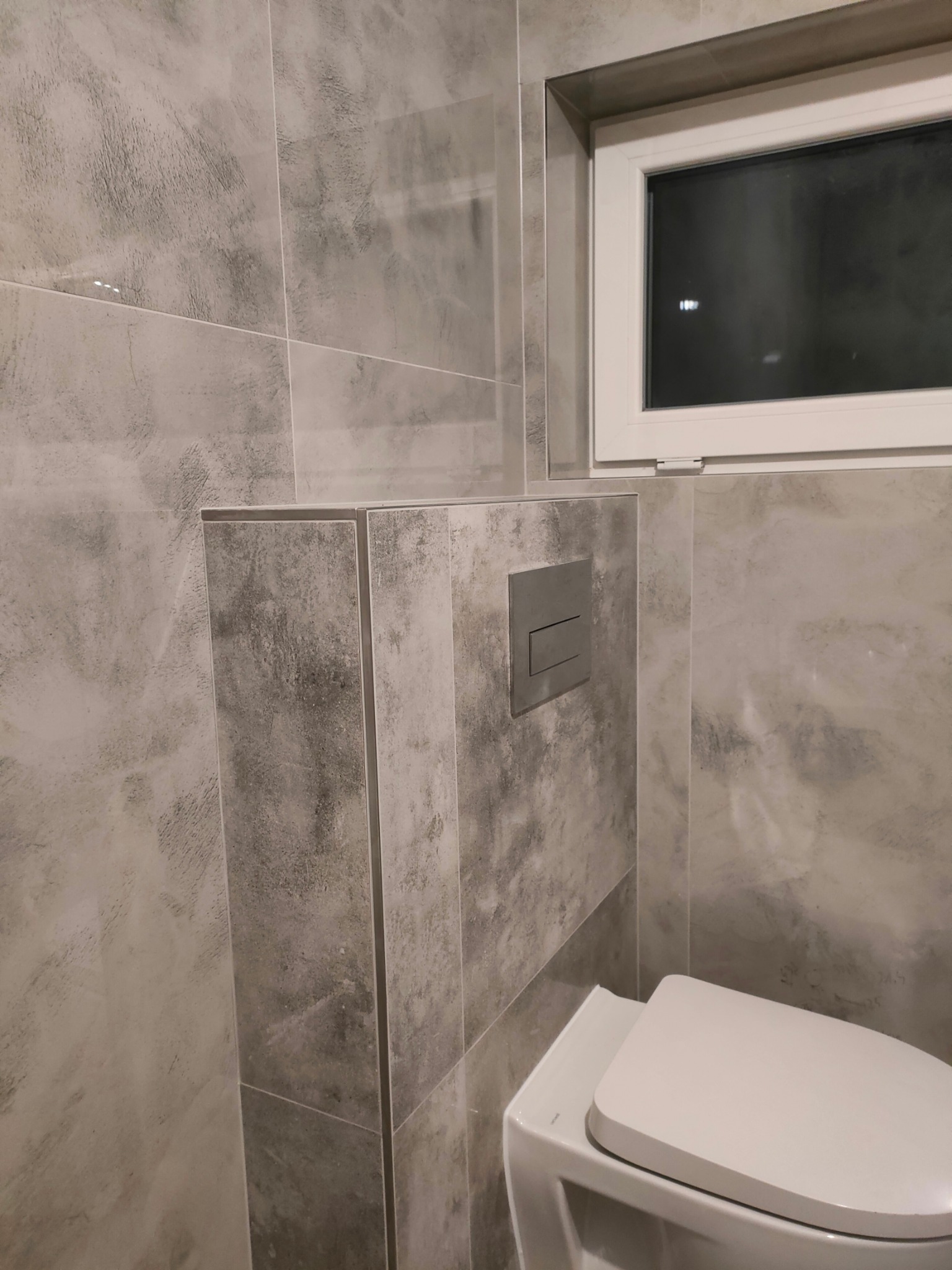 Nowoczesna łazienka wykończona szarymi płytkami imitującymi beton, z podwieszaną toaletą i minimalistycznym przyciskiem spłukującym oraz oknem w białej ramie.