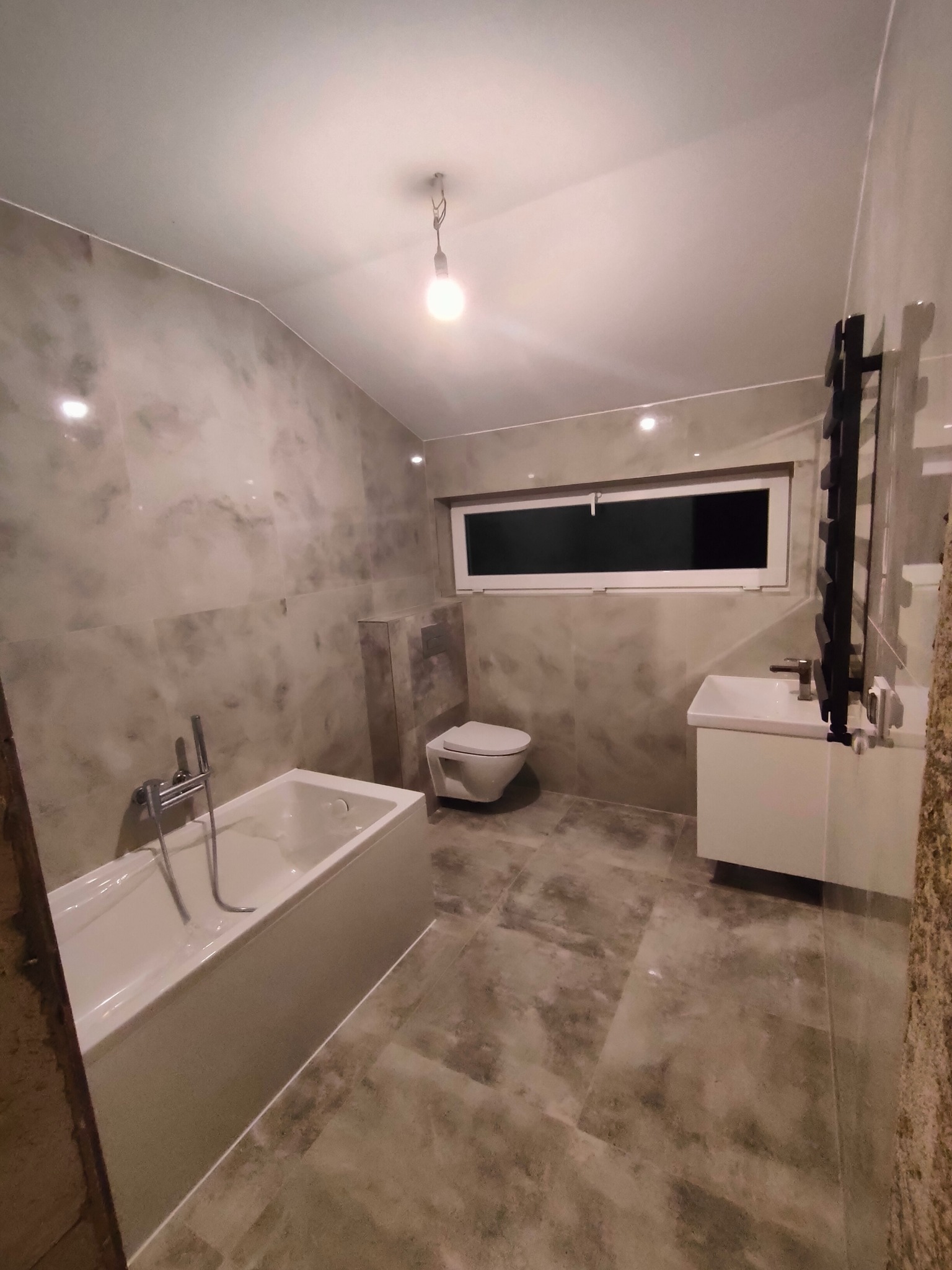 Wykończona łazienka w szarościach z wanną, umywalką z szafką i grzejnikiem drabinkowym, oświetlona pojedynczą żarówką.