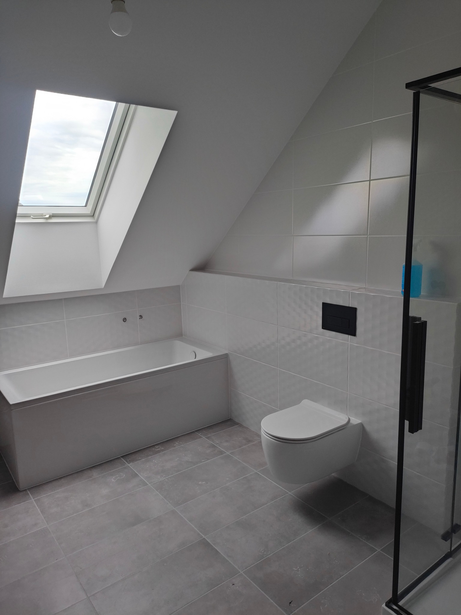 Wykończona łazienka na poddaszu z oknem dachowym, biała wanna, podwieszana toaleta i kabina prysznicowa z czarnymi profilami, szare płytki podłogowe i białe płytki ścienne z subtelnym wzorem.