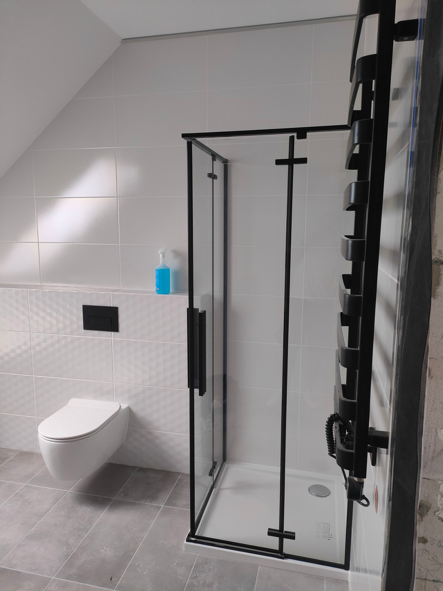 Nowoczesna łazienka na poddaszu z białą armaturą, kabiną prysznicową z czarnymi profilami i grzejnikiem drabinkowym w czarnym kolorze, białe płytki na ścianach, szare płytki na podłodze, wisząca...