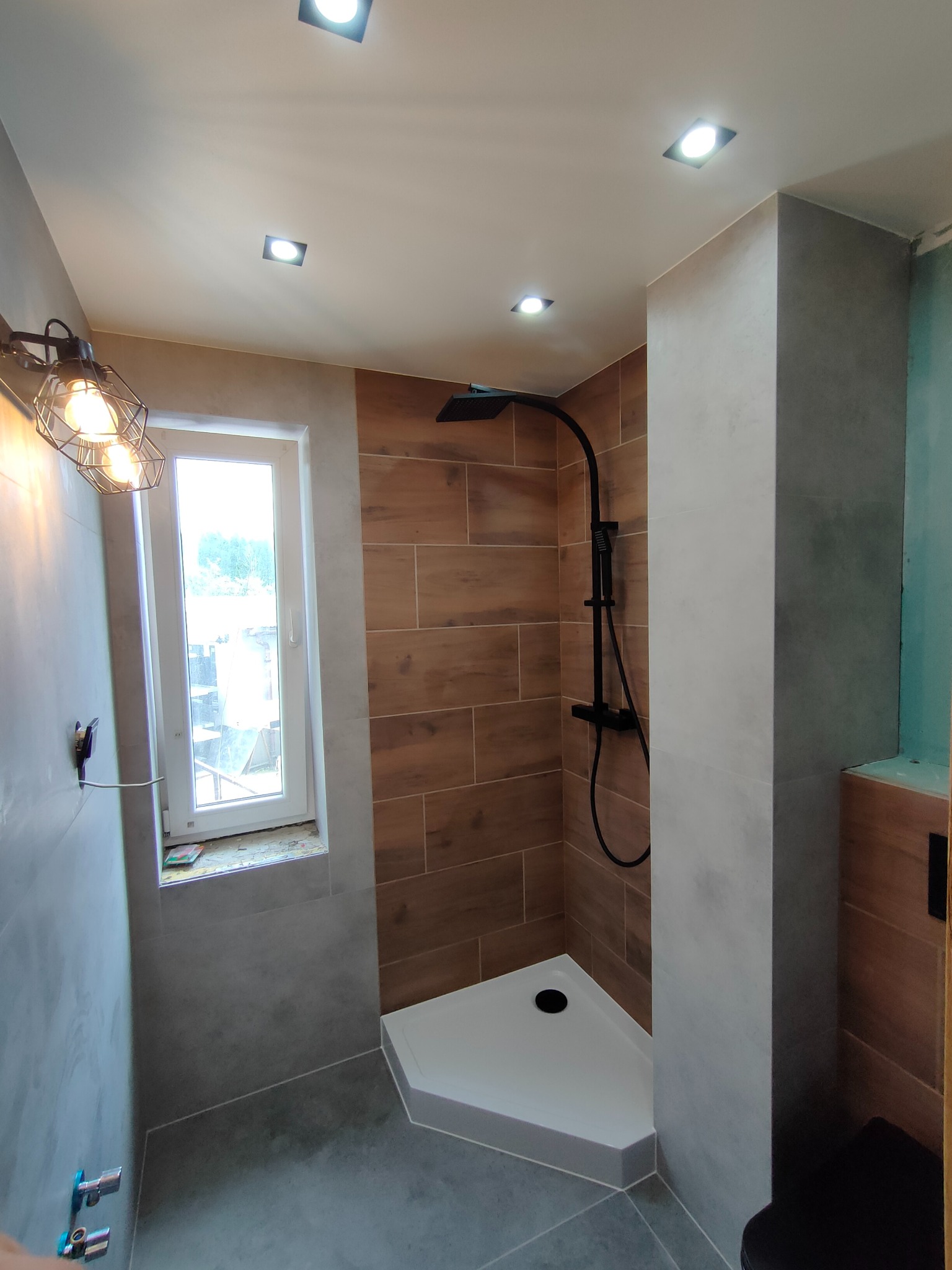 Nowoczesna kabina prysznicowa z drewnopodobnymi płytkami na ścianie, czarną armaturą i nietypowym oświetleniem przy oknie.