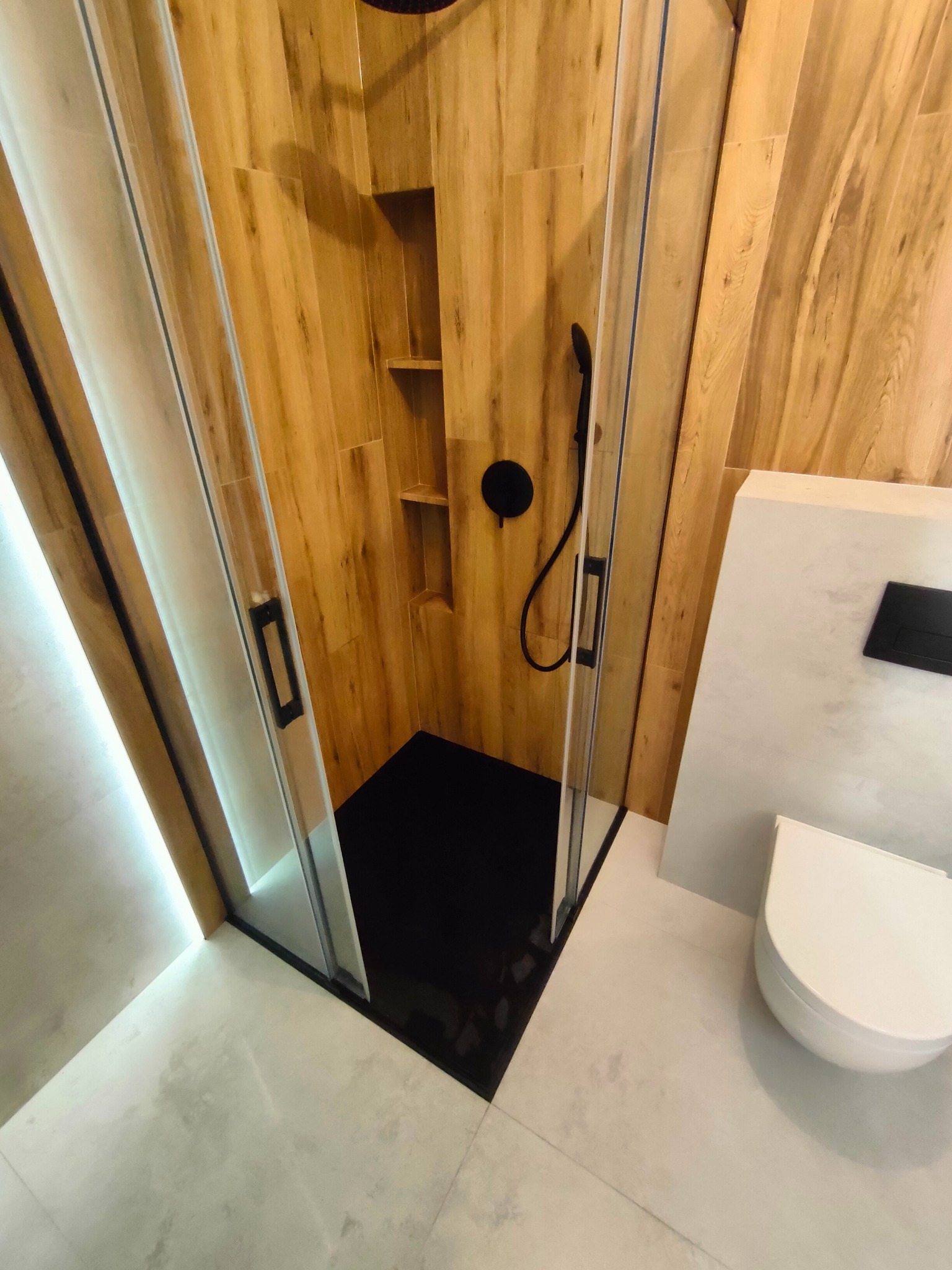 Nowoczesna łazienka z kabiną prysznicową typu walk-in, wykończona płytkami imitującymi drewno i szarym gresem, z czarną armaturą i podwieszaną toaletą.