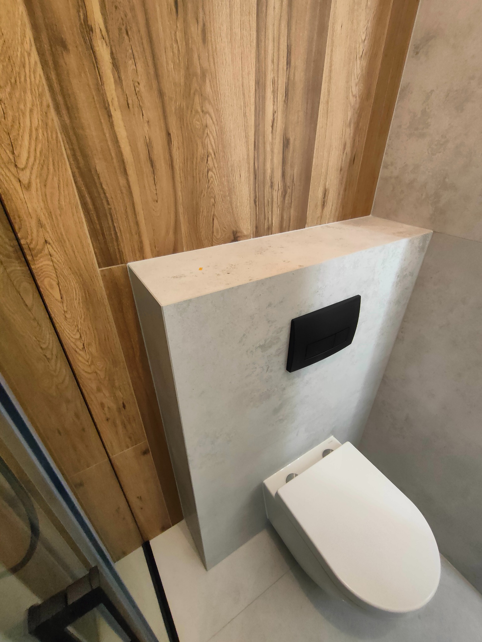 Nowoczesna toaleta podwieszana z czarnym panelem spłukującym, obudowana płytami imitującymi beton, w łazience z drewnianą okładziną ścienną