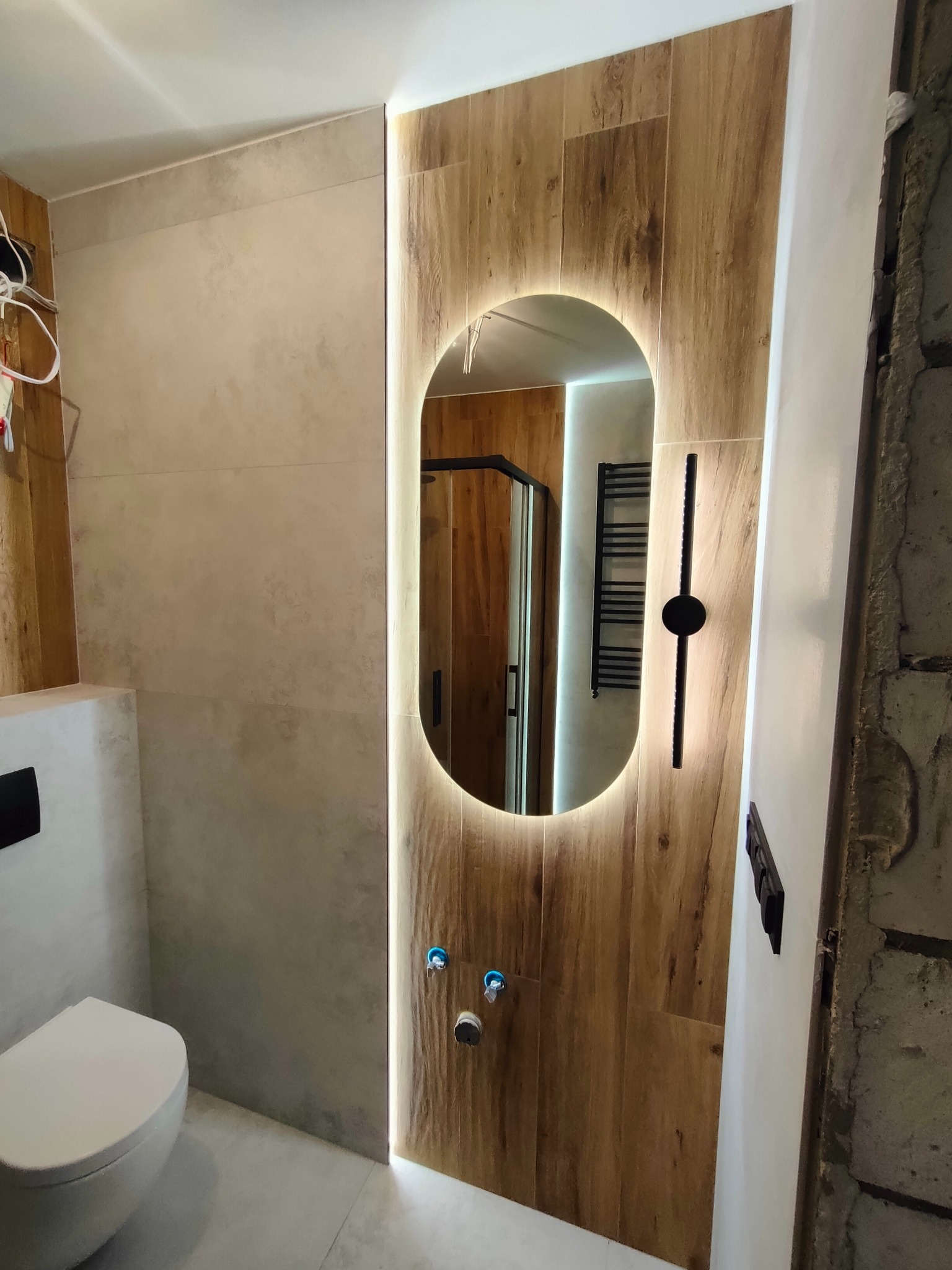 Nowoczesna łazienka z podświetlanym lustrem owalnym, drewnopodobne płytki na ścianie, wisząca toaleta i widoczny fragment kabiny prysznicowej.