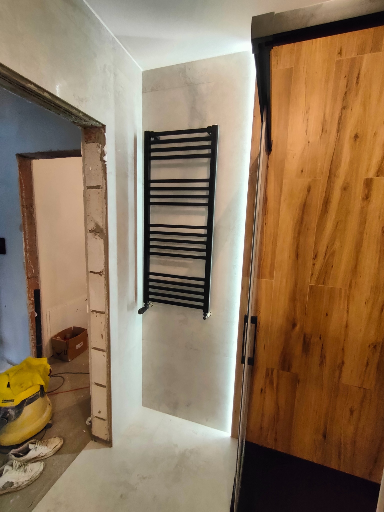 Łazienka w trakcie remontu: zamontowany czarny grzejnik drabinkowy na jasnoszarych płytkach, widoczny fragment drewnianej ściany z czarną ramą, odkurzacz i brudne buty w przejściu do innego...