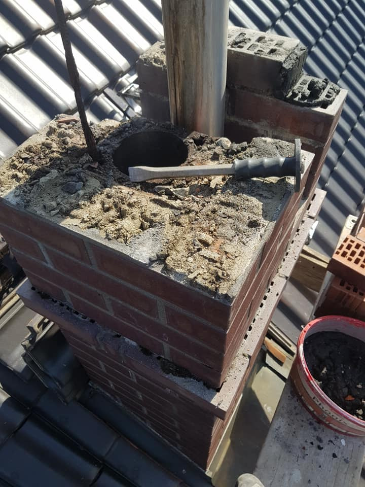 Ceglany komin na dachu w trakcie naprawy, widoczny otwór, stalowa rura i dłuto murarskie leżące na górnej powierzchni komina, częściowo uszkodzona zaprawa, dachówka w tle.