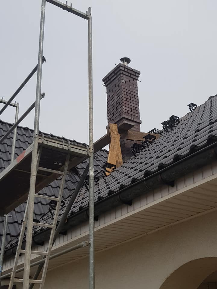 Montaż ceglanego komina na dachu z dachówki ceramicznej, widoczne rusztowanie i drewniane podpory.