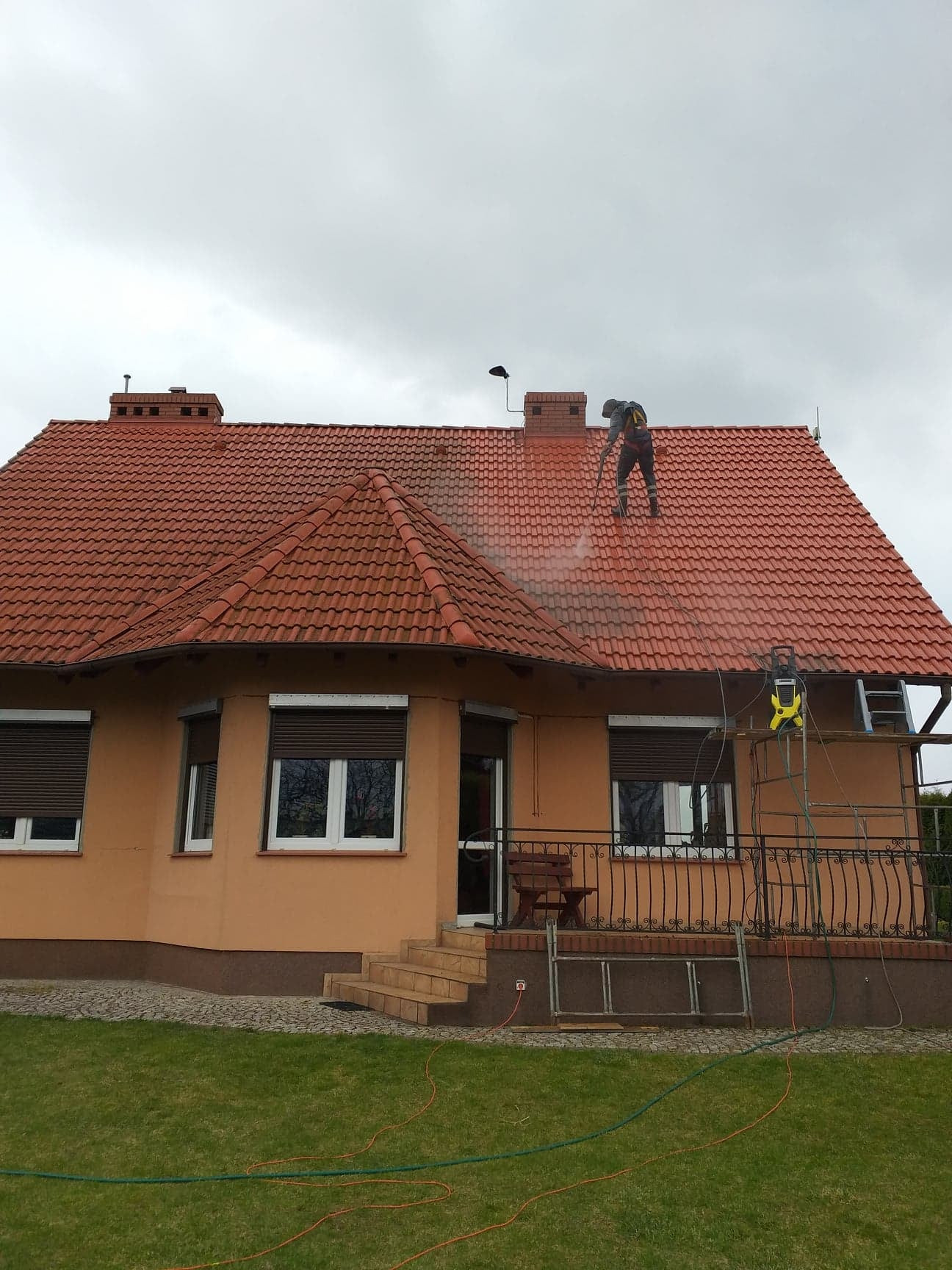 Pracownik w kombinezonie roboczym czyści czerwoną dachówkę domu jednorodzinnego za pomocą myjki ciśnieniowej, widoczna drabina i rusztowanie, pochmurne niebo w tle.