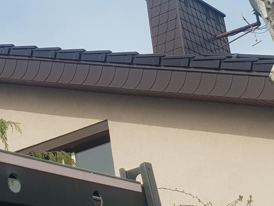 Nowy, ciemnobrązowy dach z dachówki bitumicznej w kształcie łuski, z widocznym kominem również pokrytym dachówką, antena satelitarna na kominie, jasna elewacja budynku i fragment okna.