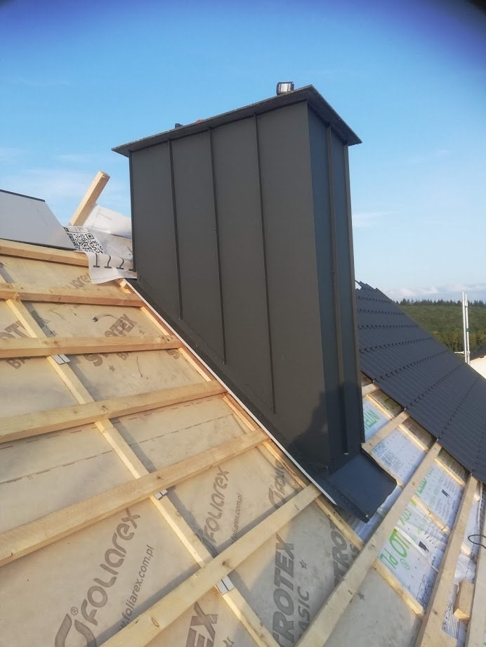 Montaż obróbki blacharskiej komina na nowym dachu, widoczna konstrukcja drewniana i membrana dachowa, w tle las i błękitne niebo.