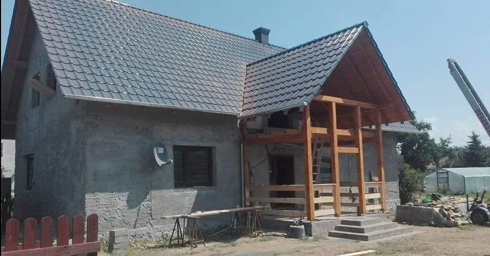 Dom w trakcie budowy z nowym szarym dachem z blachodachówki i drewnianą konstrukcją werandy, widoczna drabina i materiały budowlane.