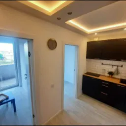 Nowoczesna kuchnia z czarnymi szafkami, drewnianym blatem i białymi kafelkami na ścianie, widok na balkon z czarnym stolikiem i tacą, oświetlenie sufitowe LED.