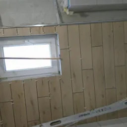 Montaż drewnopodobnych płytek ściennych wokół okna w trakcie prac, widoczne krzyżyki dystansowe i poziomica.