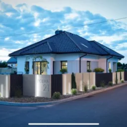 Nowoczesny dom jednorodzinny z podświetlonym ogrodzeniem z elementami betonowymi i metalowymi, ozdobiony niskimi krzewami i małymi drzewkami, widoczny z ulicy o zmierzchu.