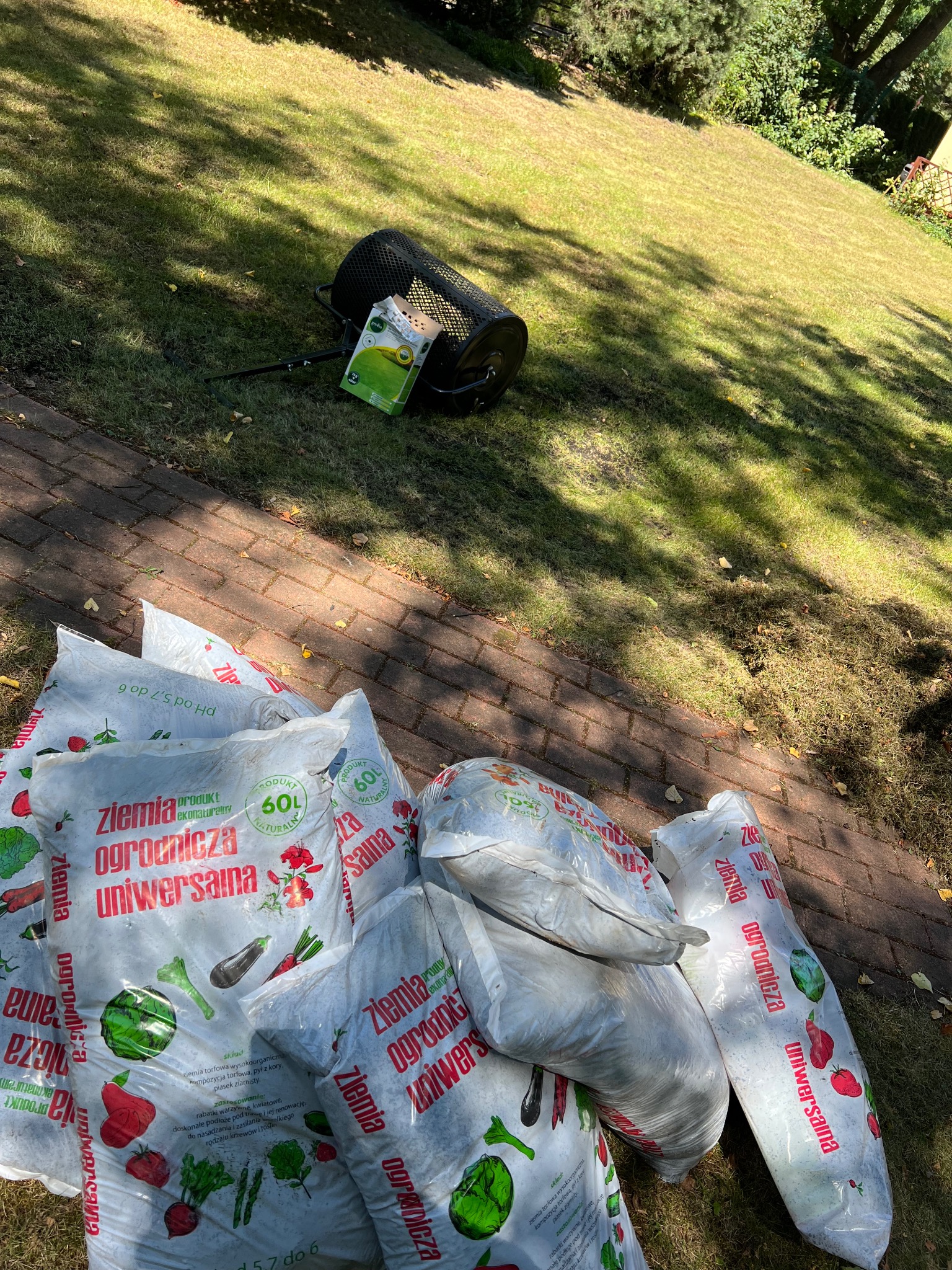 Stos worków z ziemią uniwersalną leży na kostce brukowej obok trawnika, w tle widoczny rozsiewacz do nawozu i opakowanie nasion trawy.