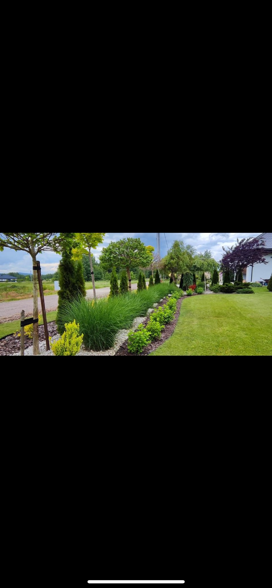 Starannie zaprojektowany i wykonany ogród z różnorodną roślinnością: drzewa formowane, tuje, trawy ozdobne, kwitnące krzewy, rabata wysypana białym kamieniem, równo przystrzyżony trawnik.