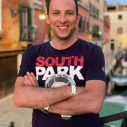 Uśmiechnięty mężczyzna w koszulce South Park, z zegarkiem na ręce i okularami na głowie, stoi z założonymi rękami na tle weneckiego kanału z budynkami w ciepłych barwach.