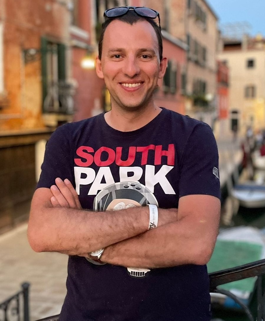 Uśmiechnięty mężczyzna w koszulce South Park, z zegarkiem na ręce i okularami na głowie, stoi z założonymi rękami na tle weneckiego kanału z budynkami w ciepłych barwach.