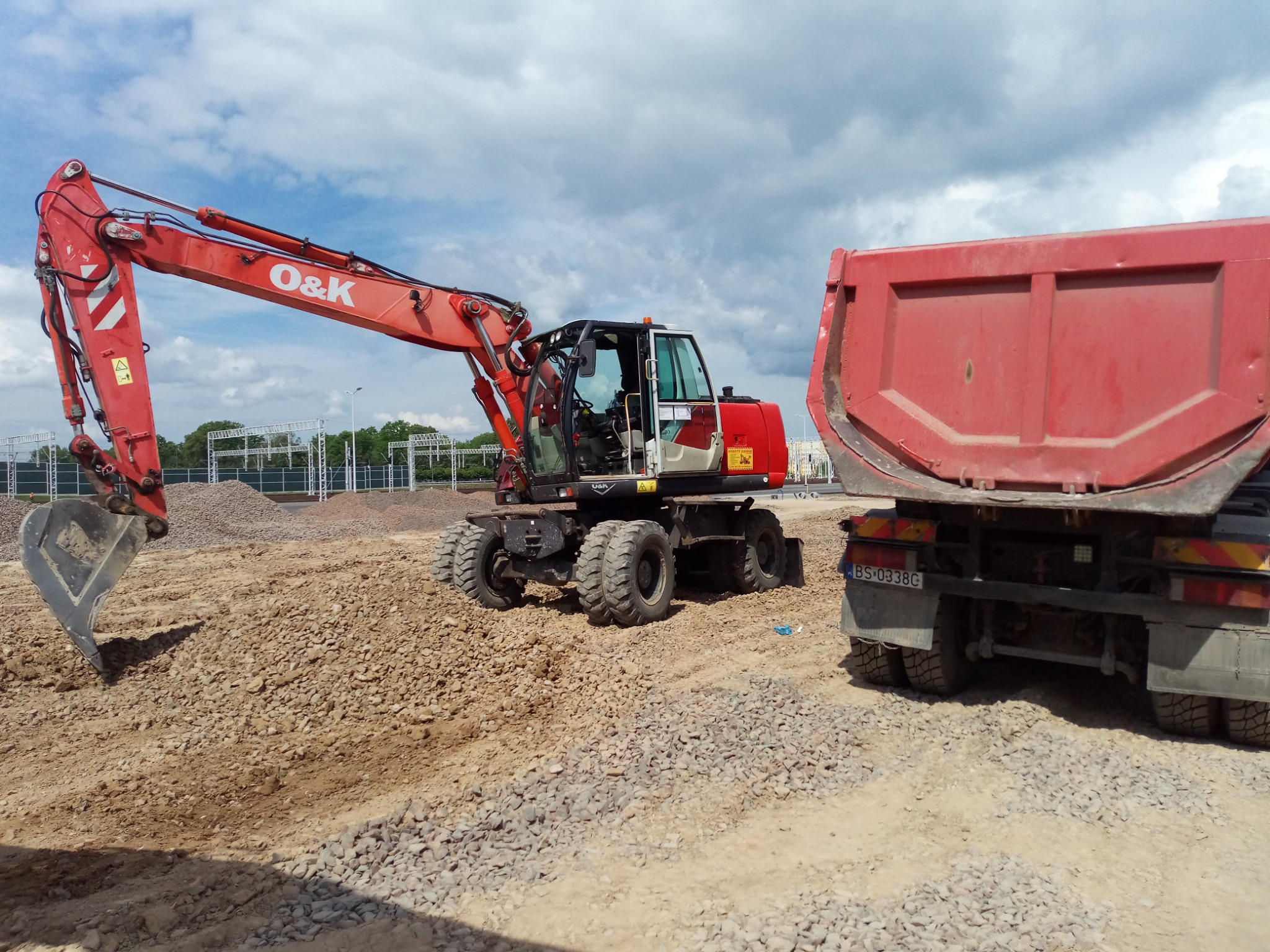 Czerwona koparka kołowa O&K z łyżką i wywrotka na placu budowy z usypanym gruzem, w tle słupy wysokiego napięcia.