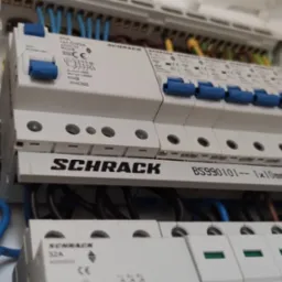 Rozdzielnica elektryczna marki Schrack z zainstalowanymi bezpiecznikami i okablowaniem w kolorach niebieskim, czarnym, żółtym i zielonym.