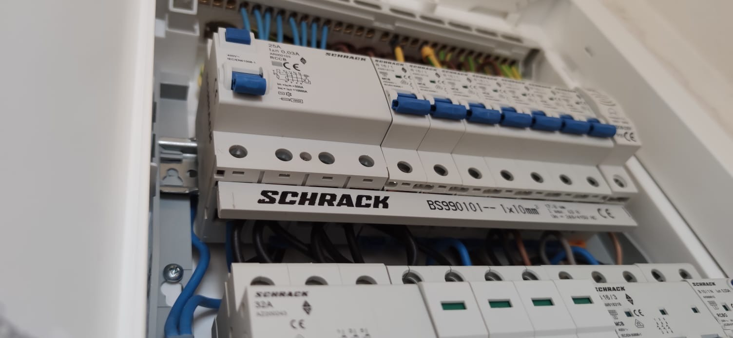 Rozdzielnica elektryczna marki Schrack z zainstalowanymi bezpiecznikami i okablowaniem w kolorach niebieskim, czarnym, żółtym i zielonym.