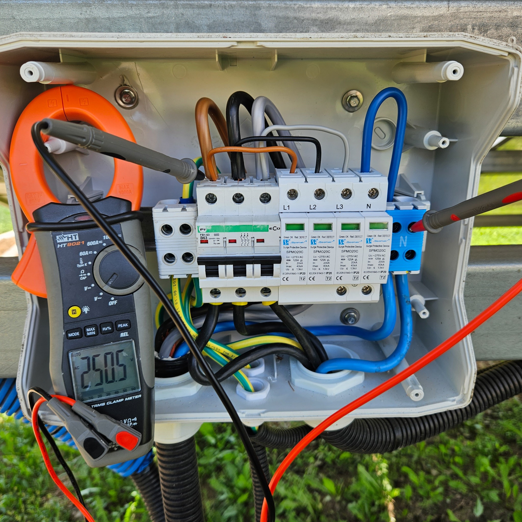 Pomiar napięcia multimetrem cęgowym HT9021 w rozdzielnicy z zabezpieczeniami przepięciowymi Spamel SPMO20C, widoczne kolorowe przewody instalacji elektrycznej.