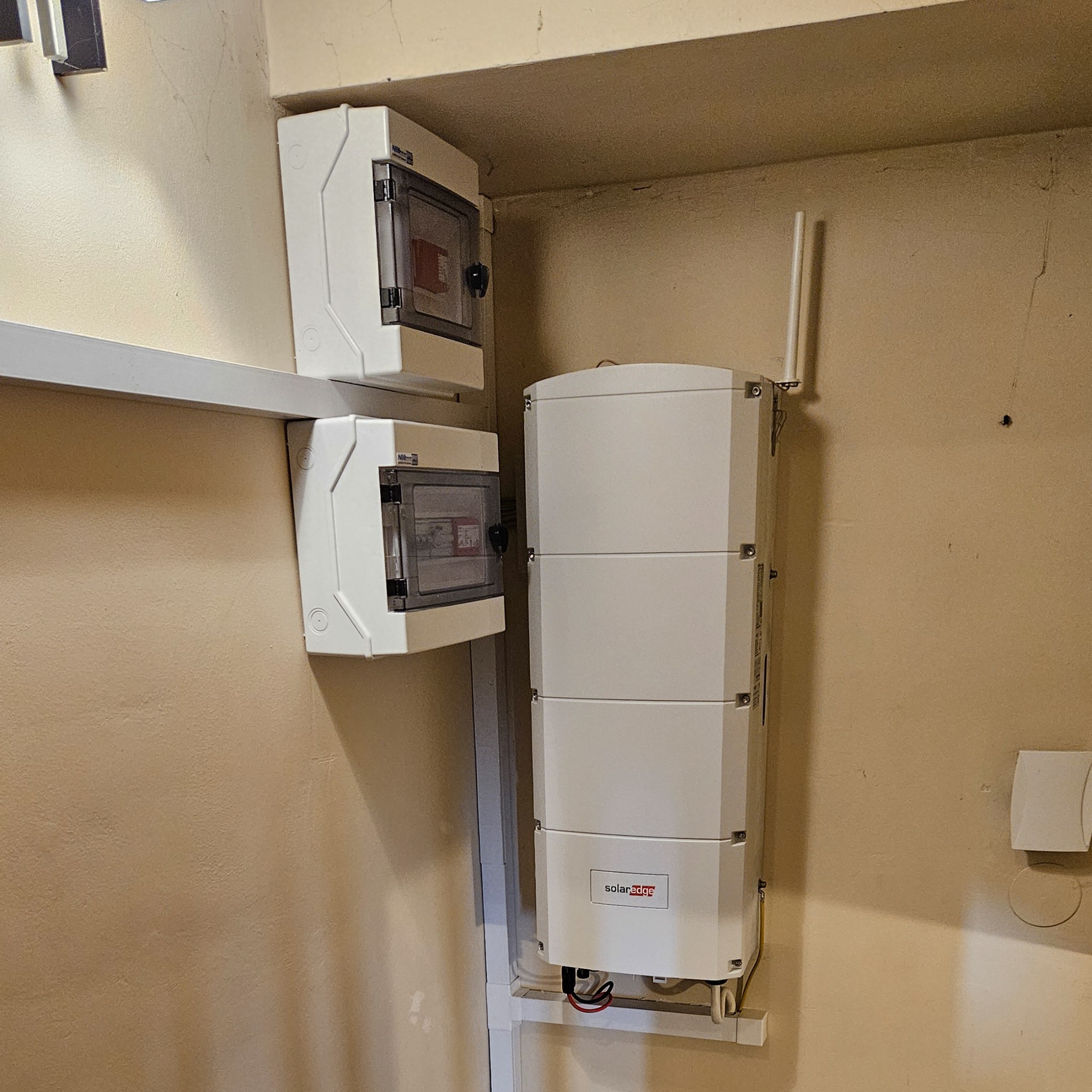 Białe skrzynki elektryczne i modułowa obudowa z anteną, prawdopodobnie falownik, zamontowane na ścianie wewnątrz budynku, widoczne przewody.