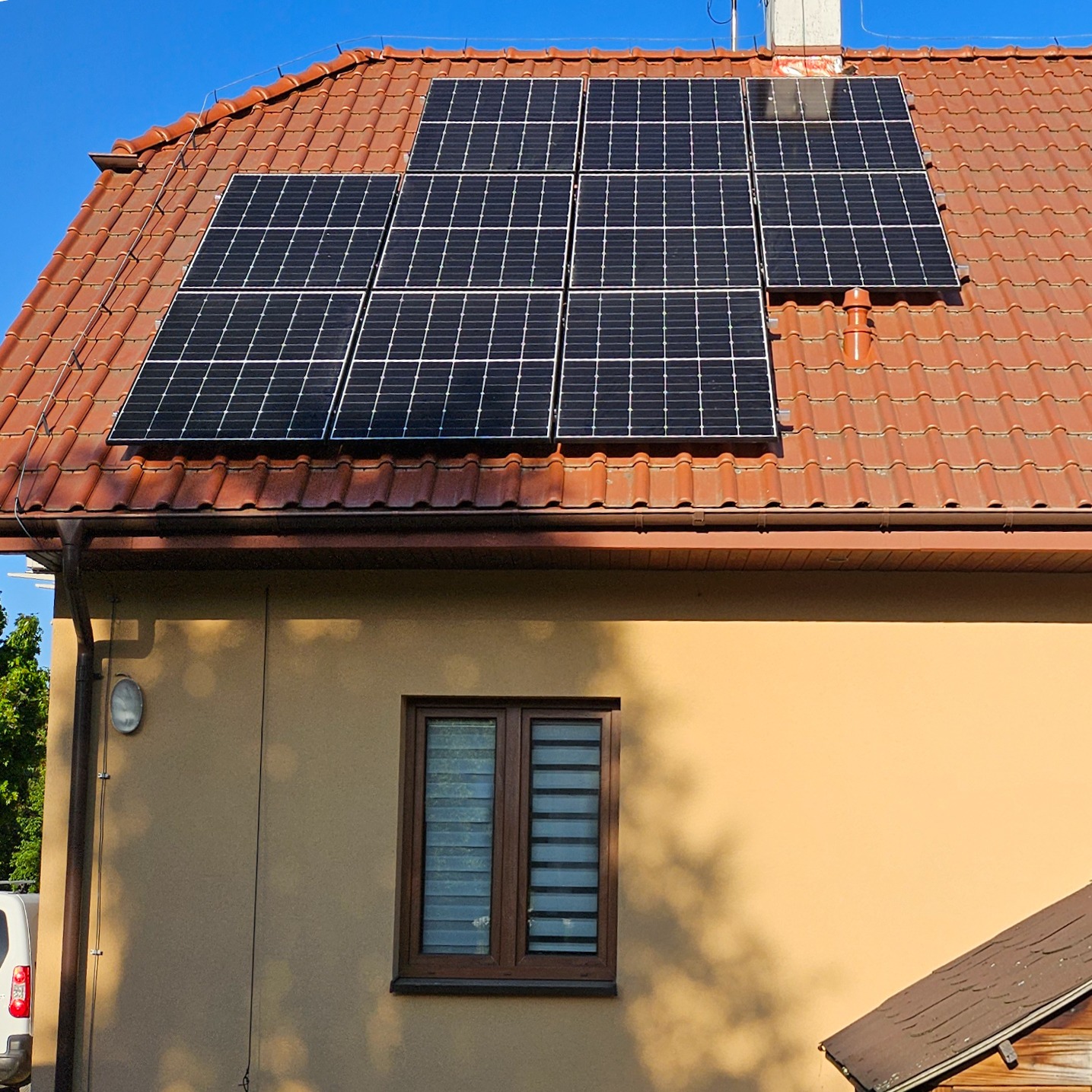 Panele słoneczne na dachu domu z brązową dachówką i beżową elewacją w słoneczny dzień.