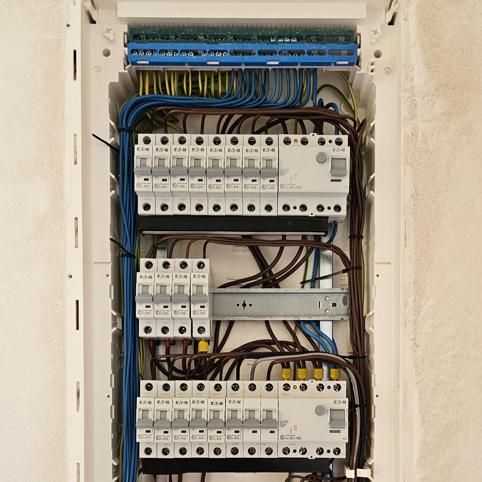 Otwarta skrzynka elektryczna z zainstalowanymi bezpiecznikami EATON, widoczne różnokolorowe przewody i listwy zaciskowe, zamontowana na nierównej, pomalowanej ścianie.