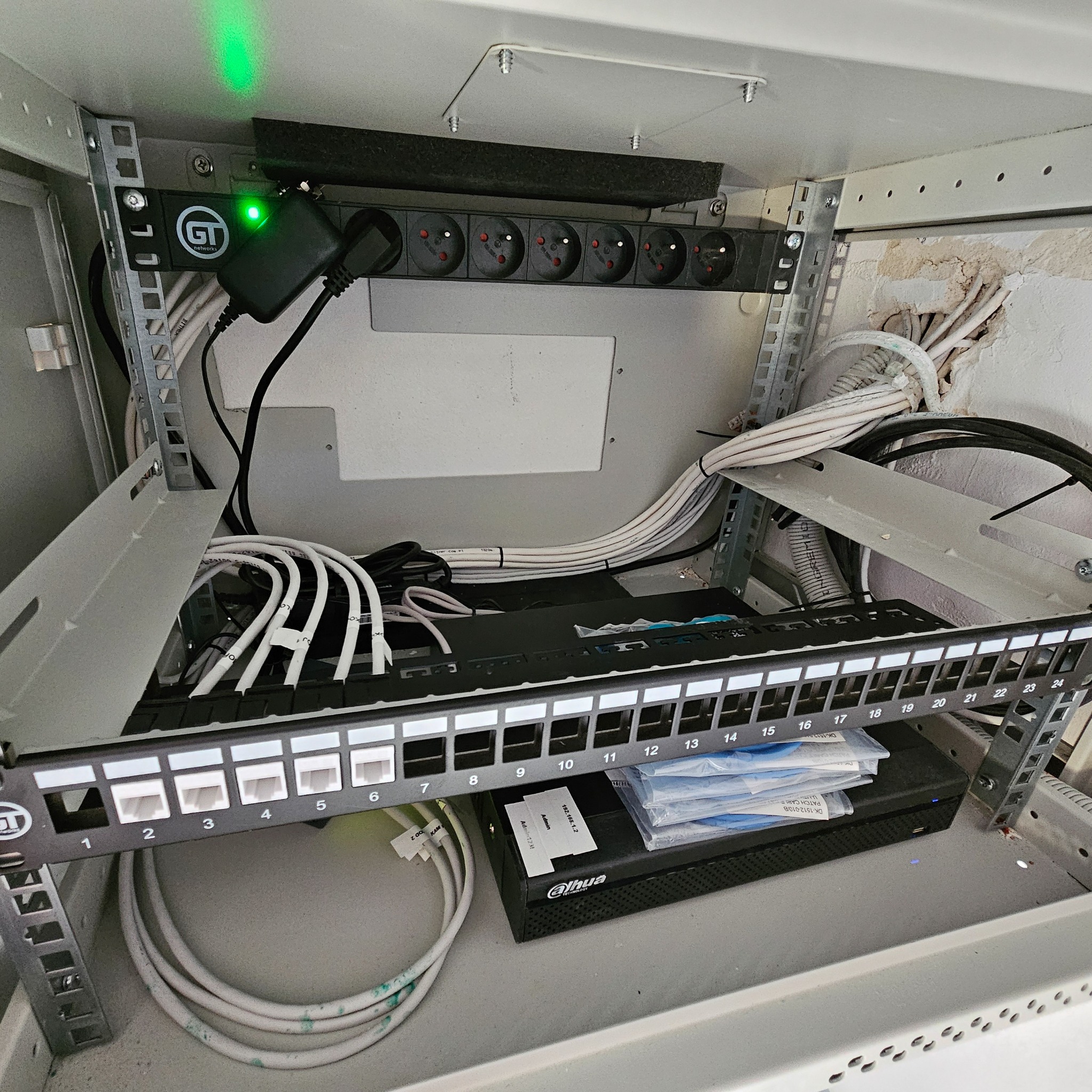 Szafa rack z patch panelem, listwą zasilającą z gniazdami europejskimi, zasilaczem GT Networks ze świecącą zieloną diodą, okablowaniem strukturalnym i rejestratorem Dahua z naklejką konfiguracyjną.