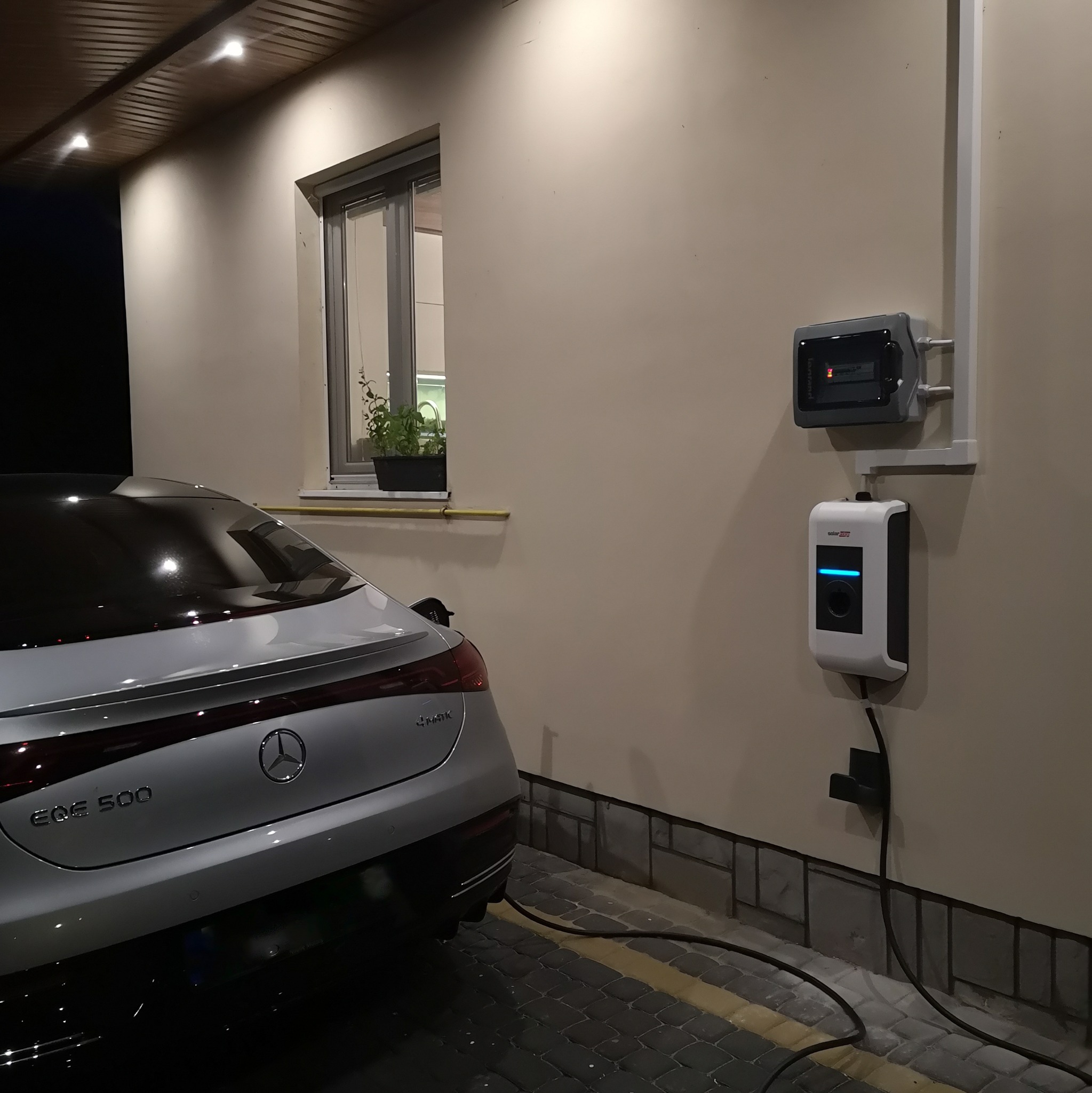 Biały samochód elektryczny Mercedes EQS 500 podłączony do stacji ładowania SolarEdge na ścianie budynku, widoczna skrzynka elektryczna i kabel zasilający.