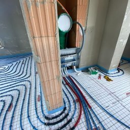 HYD-GUT - Instalacja ogrzewania podłogowego w trakcie montażu, widoczne rury w kolorze niebieskim i czerwonym, rozdzielacz, oraz fragment surowego muru z cegły.
