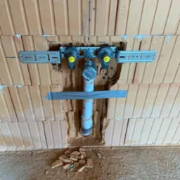Fragment instalacji hydraulicznej w trakcie montażu w ścianie z cegły, widoczne mocowania, rura kanalizacyjna zabezpieczona taśmą, drobne gruzowisko na podłodze.