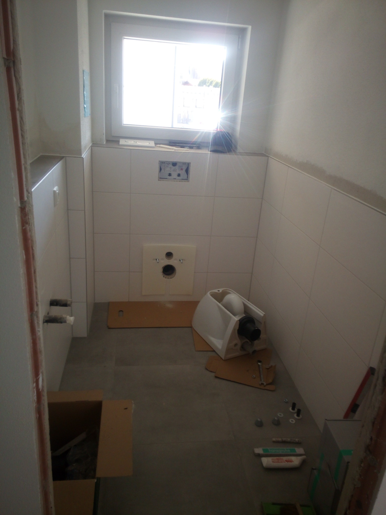 Wnętrze łazienki w trakcie remontu, widoczna instalacja podtynkowego WC ze stelażem, białe płytki na ścianach, kartony i narzędzia na podłodze, okno z widokiem na zewnątrz.