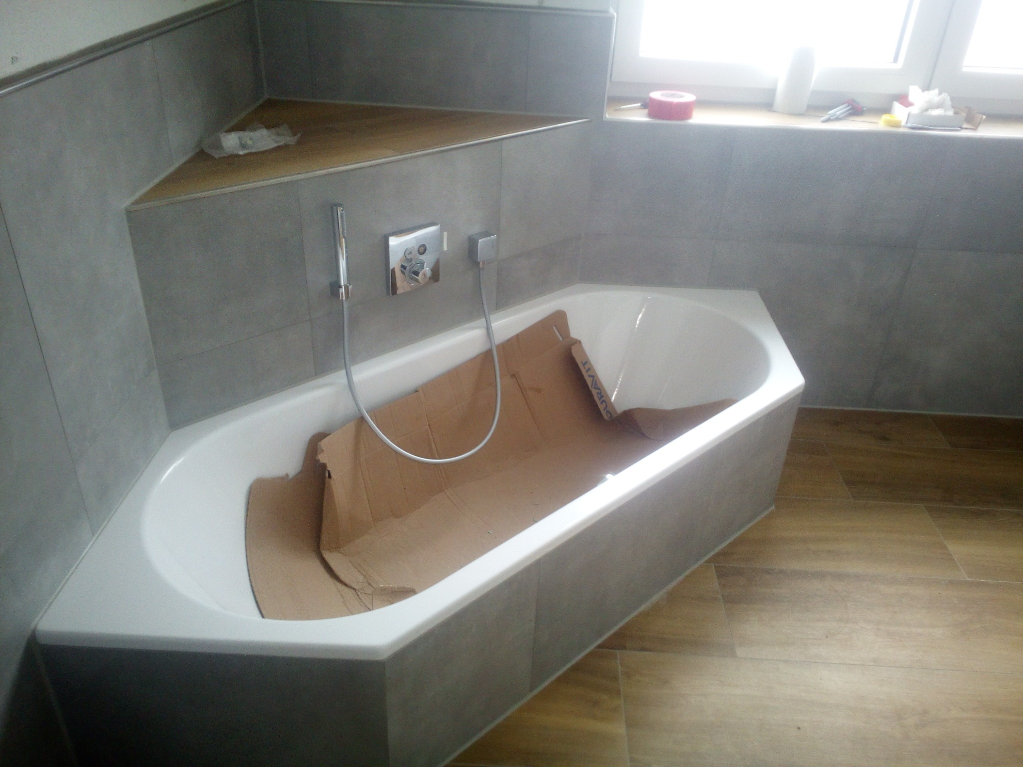 Narożna wanna akrylowa zabezpieczona kartonem podczas prac wykończeniowych w łazience z szarą glazurą i drewnopodobną podłogą, widoczna bateria natryskowa i okno w tle.