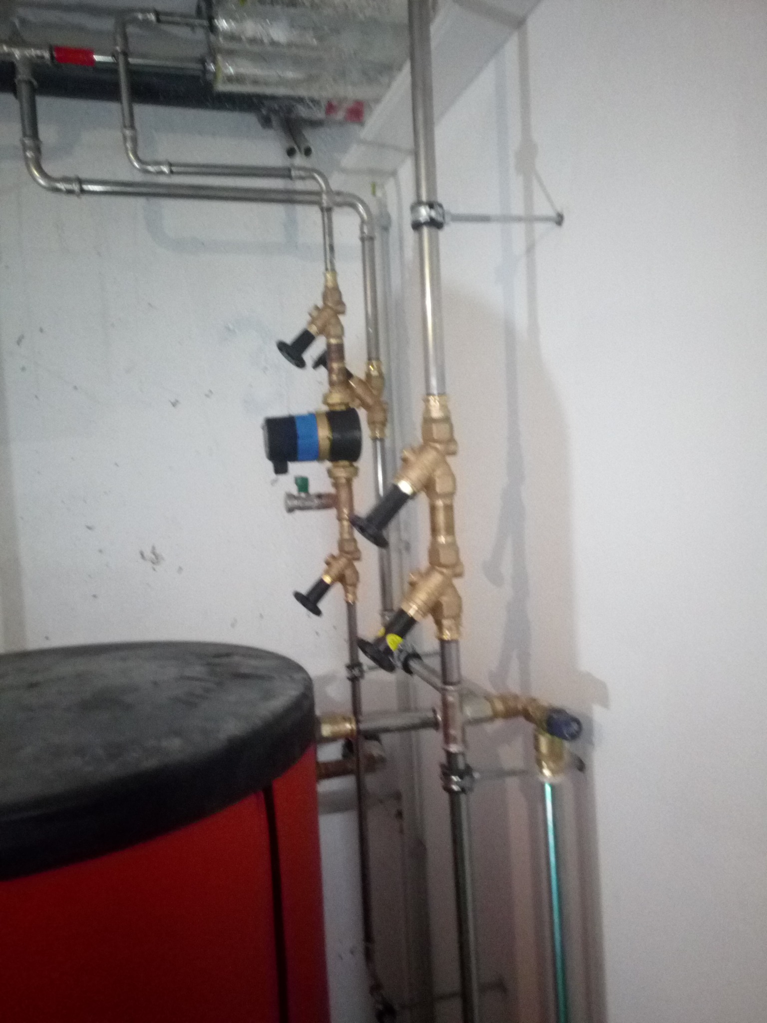 Czerwony zasobnik ciepłej wody użytkowej z podłączonymi rurami miedzianymi i stalowymi, zawory regulacyjne z czarnymi pokrętłami, instalacja w pomieszczeniu technicznym o surowych ścianach.