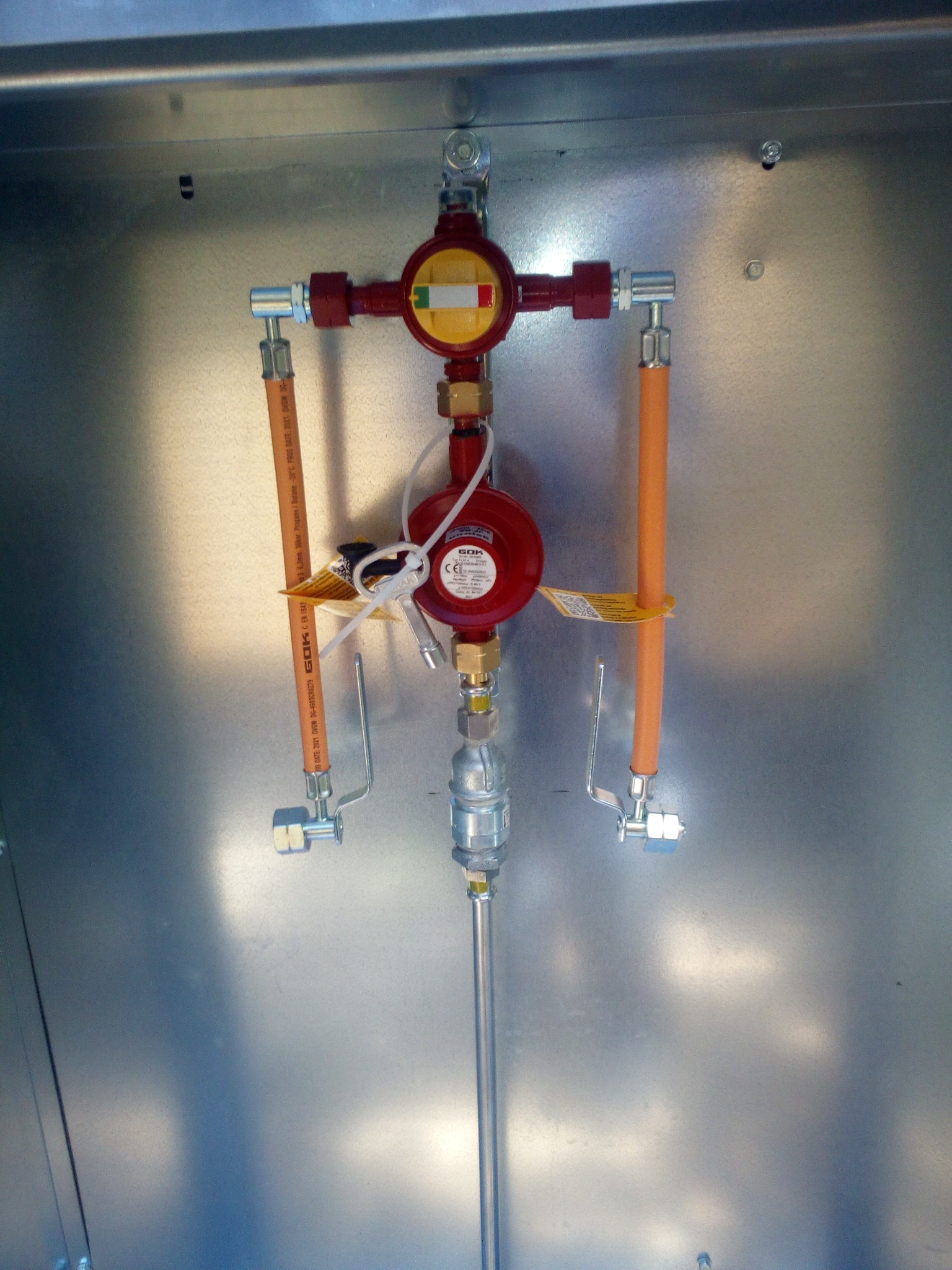 Zmontowany reduktor ciśnienia gazu z przewodami w metalowej obudowie, widoczne oznaczenia producenta GOK i certyfikaty na przewodach gazowych.