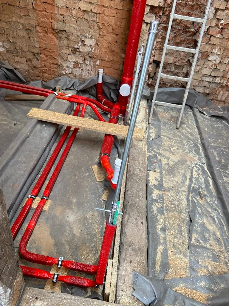 Czerwone rury ogrzewania podłogowego w trakcie montażu, przymocowane do drewnianej konstrukcji, widoczne elementy mocujące, szara folia izolacyjna, ceglana ściana w tle i aluminiowa drabina oparta...