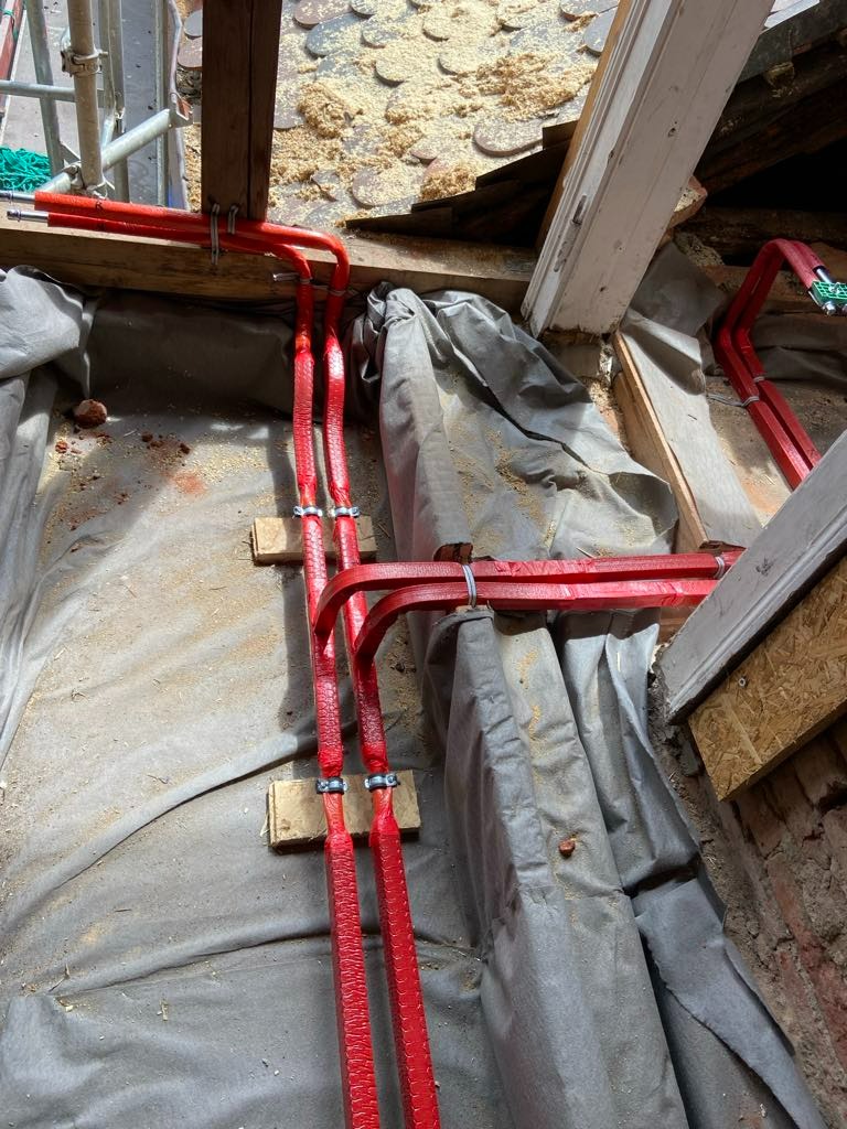 Czerwone rury instalacji grzewczej, zabezpieczone folią budowlaną, montowane pod podłogą w trakcie remontu, widoczne drewniane elementy konstrukcyjne i ocieplenie stropu.