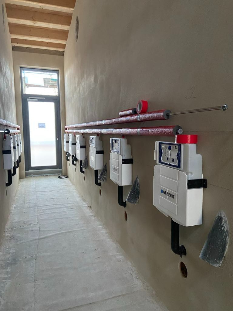 Rząd podtynkowych stelaży WC marki Geberit zamontowanych w surowym stanie budynku, rury w izolacji termicznej z czerwoną taśmą, widoczne otwory w ścianie i podłodze, brak wykończenia ścian i podłóg.