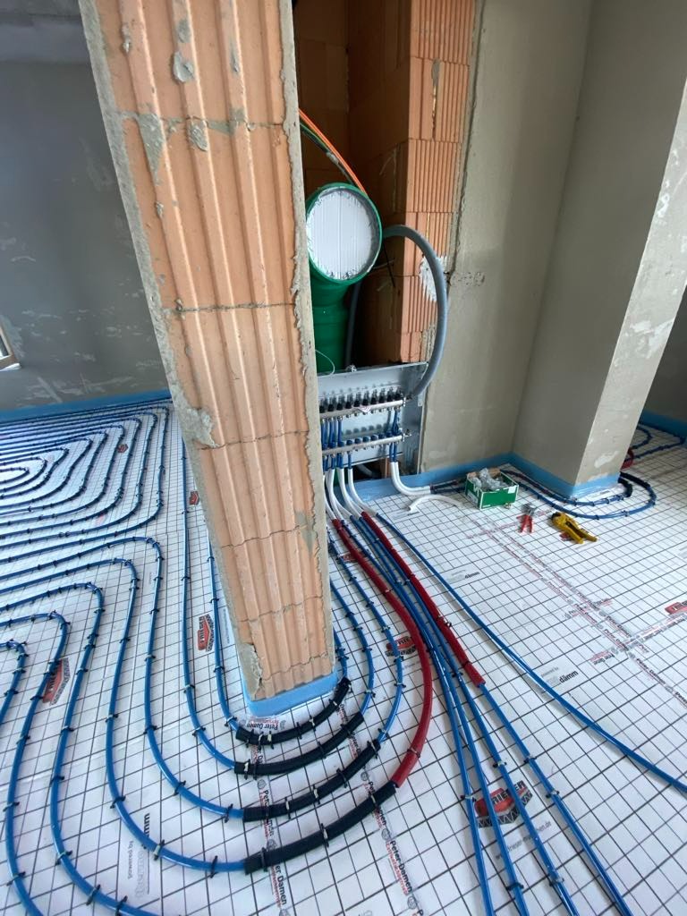 Instalacja ogrzewania podłogowego w trakcie montażu, widoczne rury w kolorze niebieskim i czerwonym, rozdzielacz, oraz fragment surowego muru z cegły.
