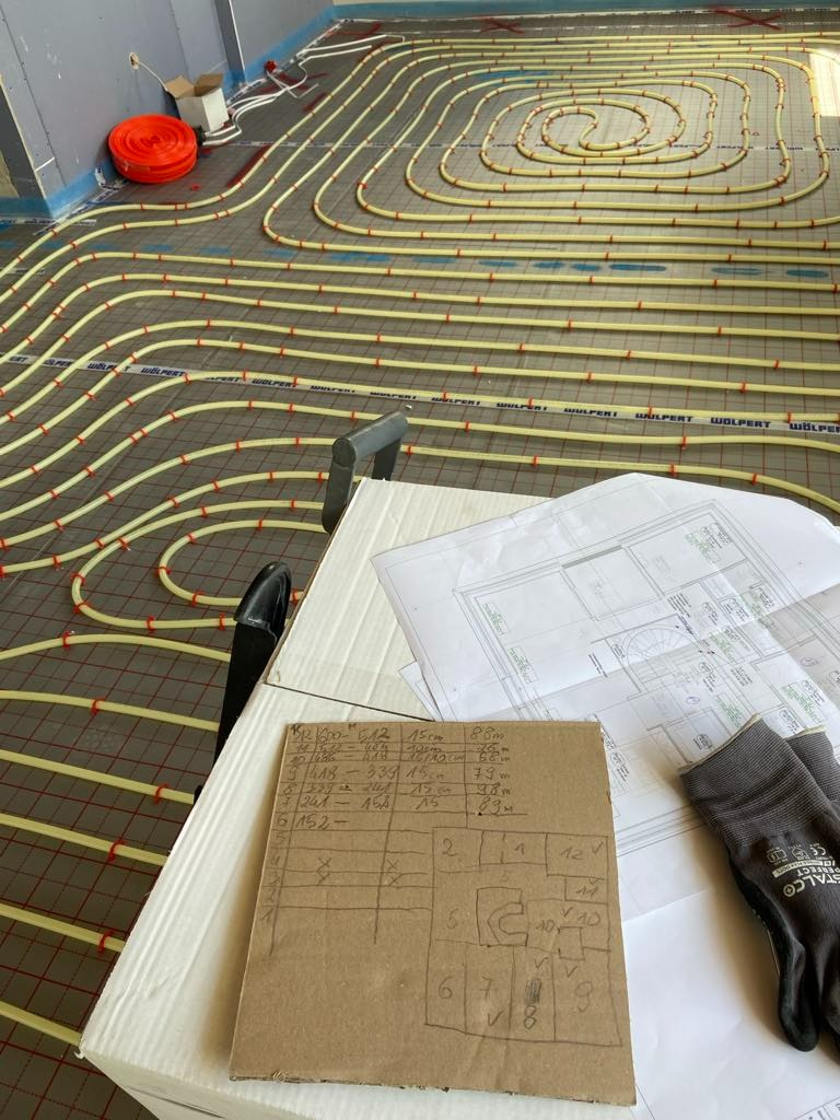 Żółte rurki instalacji grzewczej ułożone spiralnie na podłodze z siatką, obok plany instalacji, notatki na kartonie i rękawice robocze.