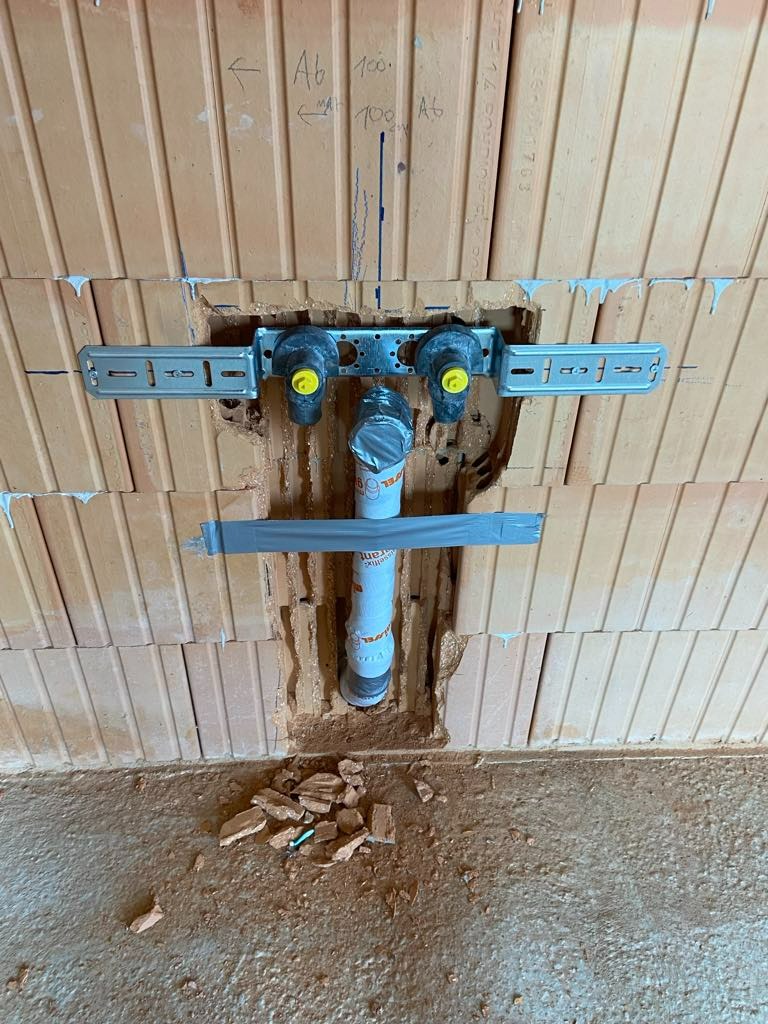 Fragment instalacji hydraulicznej w trakcie montażu w ścianie z cegły, widoczne mocowania, rura kanalizacyjna zabezpieczona taśmą, drobne gruzowisko na podłodze.