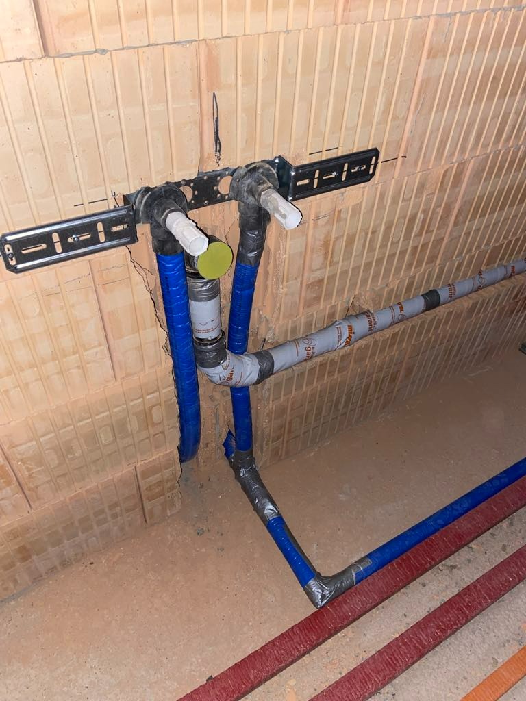 Fragment instalacji wodnej z niebieskimi rurami przymocowanymi do ściany z cegły, widoczne łączenia owinięte taśmą izolacyjną.