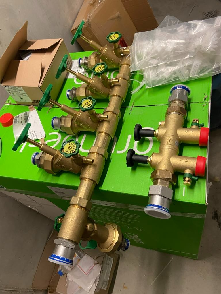 Zestaw mosiężnych zaworów i złączek hydraulicznych z zielonymi i czarnymi pokrętłami, ułożony na zielonym kartonie z logo producenta.