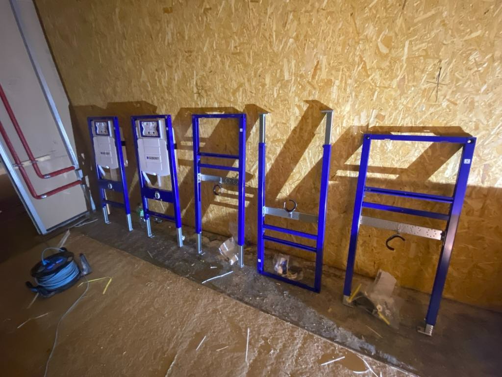 Rzędy niebieskich stelaży podtynkowych Geberit ustawione na podłodze w surowym wnętrzu z boazerią, widoczny zwój kabla i fragment instalacji rurowej.