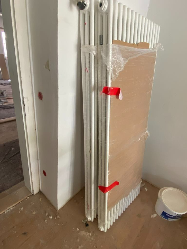 Nowe, zapakowane grzejniki żeberkowe, zabezpieczone folią i taśmą, stoją oparte o ścianę w trakcie remontu, obok widoczne wiadro z farbą.