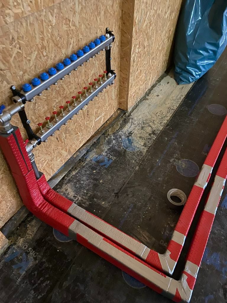 Rozdzielacz do instalacji grzewczej z licznymi wyjściami, zamontowany na ścianie z płyty OSB, z widocznymi rurami zabezpieczonymi czerwoną izolacją i srebrną taśmą, ułożonymi na czarnej folii...
