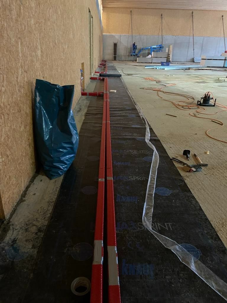 Czerwone rury grzewcze ułożone na czarnej folii izolacyjnej na podłodze w trakcie montażu, widoczny worek na śmieci, narzędzia i podnośnik w tle.